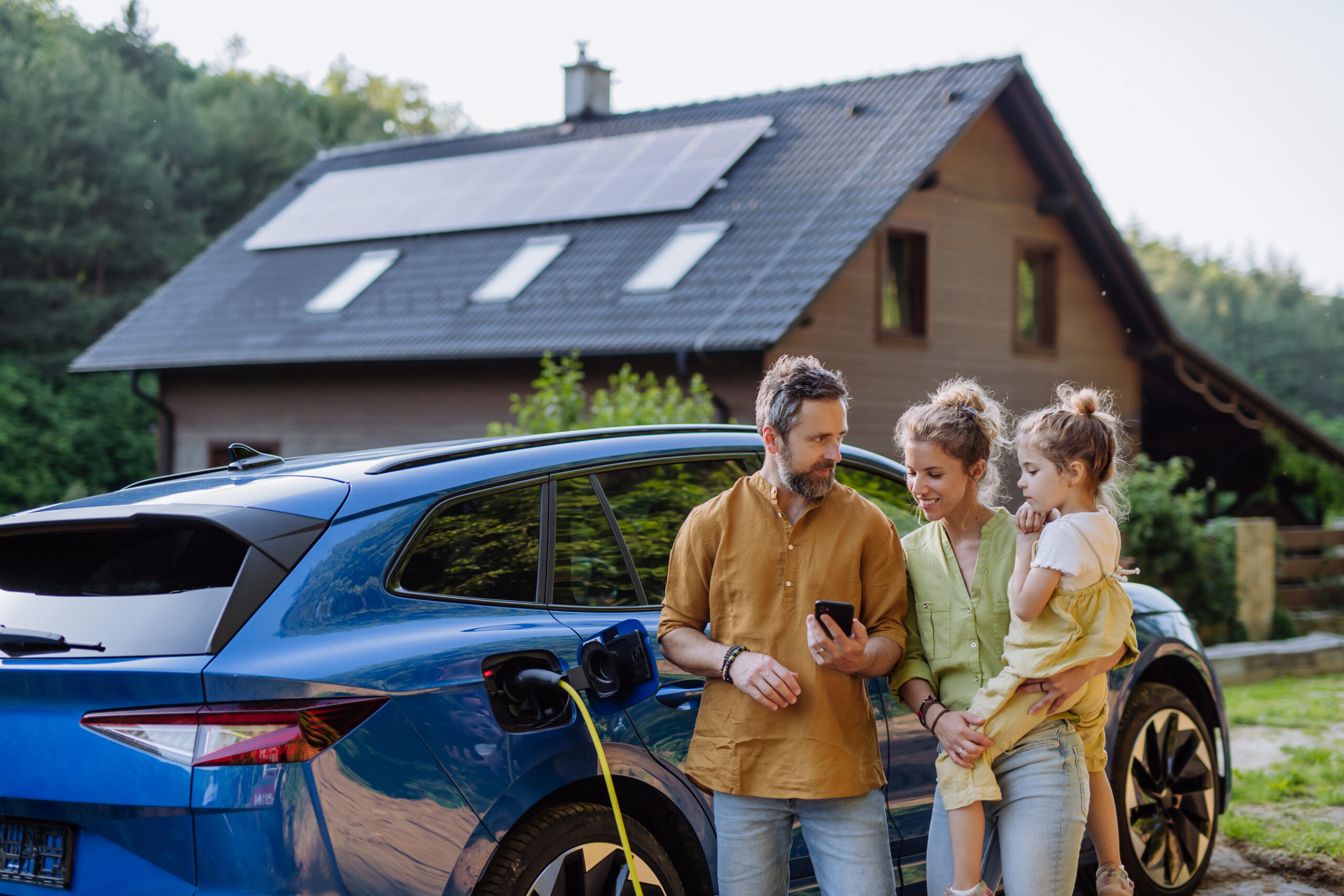 Family with little daughter standing in front of their house with solar panels on the roof, having electric car.